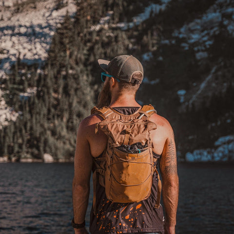 Hiker wearing the brown Adventure Pack 12L by a mountain lake, back view